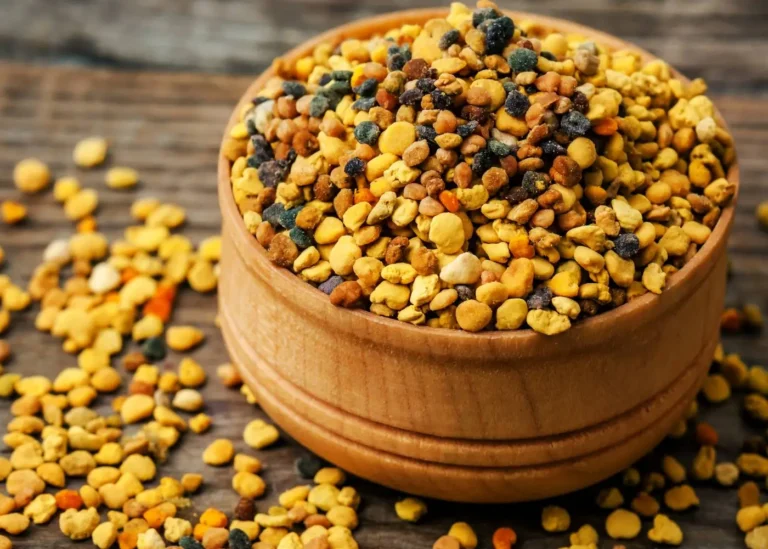 Na zdjęciu znajduje się drewniana miseczka pełna żółtych, pomarańczowych i brązowych granulek, które wyglądają na pyłek pszczeli. Pyłek jest umieszczony na drewnianym, strukturalnym tle, a część z niego wysypała się poza miseczkę. Ten naturalny produkt jest popularnym suplementem diety, cenionym za swoje wartości odżywcze. Granulki mają różne odcienie i wielkości, co świadczy o ich naturalnym pochodzeniu.