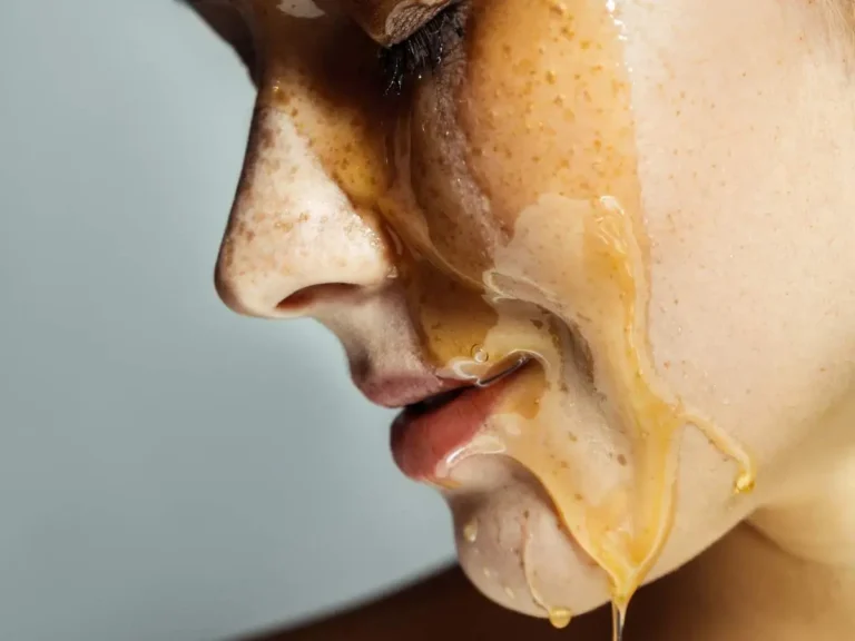 Na zdjęciu przedstawiona jest zbliżenie fragmentu twarzy osoby, na której widoczna jest maseczka z miodu. **Główne elementy zdjęcia:** - **Twarz**: Widać nos, usta oraz część policzka osoby. Skóra jest lekko zarumieniona i ma naturalną teksturę. Na twarzy widać piegi. - **Miód**: Maseczka z miodu jest gęsta i płynie w dół twarzy, tworząc lśniące, złote smugi. Miód wygląda na lepki i jest równomiernie rozprowadzony na skórze. - **Tło**: Tło zdjęcia jest neutralne, najprawdopodobniej szare, co podkreśla szczegóły twarzy i maseczki. Zdjęcie ma charakter kosmetyczny, ukazując naturalny zabieg pielęgnacyjny z użyciem miodu.