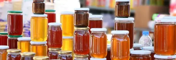 Rzędy słoików z różnymi rodzajami miodu ustawione na targowym stoisku – od jasnozłotych po ciemnoambrowe, każdy z unikalnym kolorem i konsystencją. W tle rozmyte wnętrze hali targowej. Obraz symbolizuje bogactwo i różnorodność produktów pszczelich, które można promować na stronie – od klasycznych miodów po nowoczesne wyroby naturalne.